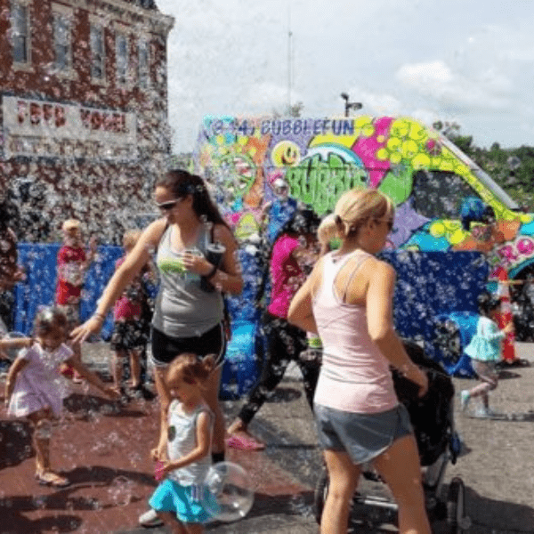 Kids and parents walking around KidsFest with bubbles in the air and tie-dye van in the background.